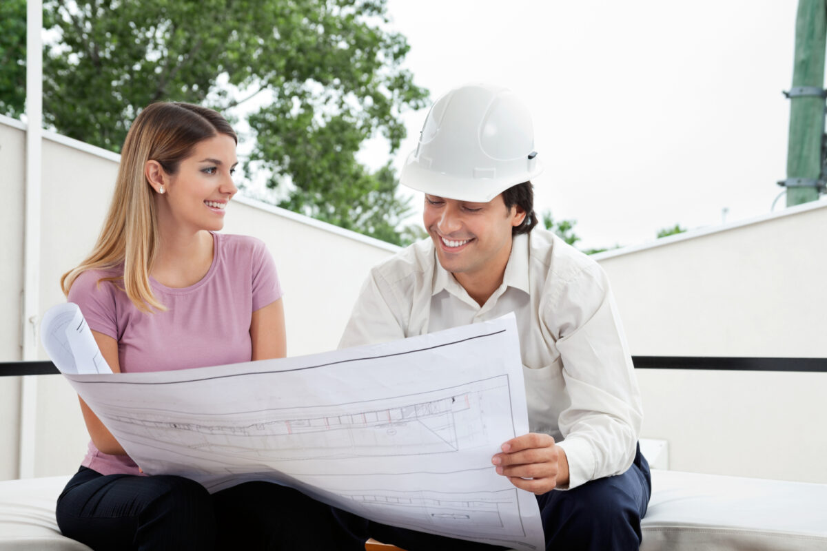 A contractor looks over a blueprint with a customer for a home renovation project. The contractor is wearing a white shirt with a white construction helmet. The customer is wearing a light purple shirt.