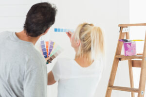 A couple look at paint swatches as they hold it up and compare it with a white wall in their home.