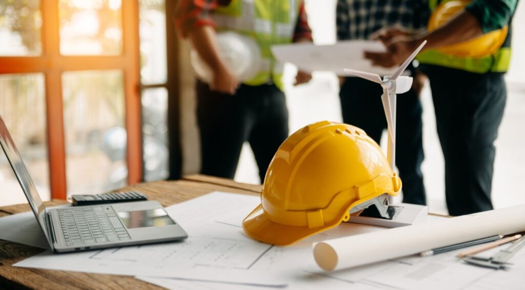 A construction helmet sits on a table with a blueprint and a laptop.